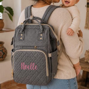 以下が含まれることがあります： ピンク色の「Noelle」の刺繍が入ったグレーのキルティングおむつバッグ。複数のコンパートメント、上部のハンドル、調節可能なショルダーストラップを備えています。ゴールドトーンのジッパーと金具。実用的でスタイリッシュな親御さんのためのアクセサリー。
