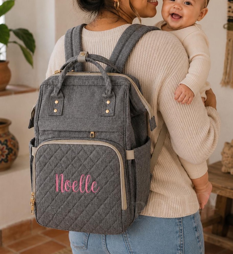 以下が含まれることがあります： ピンク色の「Noelle」の刺繍が入ったグレーのキルティングおむつバッグ。複数のコンパートメント、上部のハンドル、調節可能なショルダーストラップを備えています。ゴールドトーンのジッパーと金具。実用的でスタイリッシュな親御さんのためのアクセサリー。