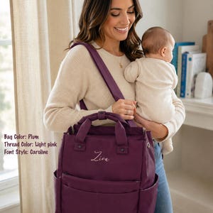 May include: Plum-colored diaper bag with light pink embroidered name "Ziva". The bag has multiple pockets and a top handle, and is carried over the shoulder. The bag is a stylish accessory for parents.