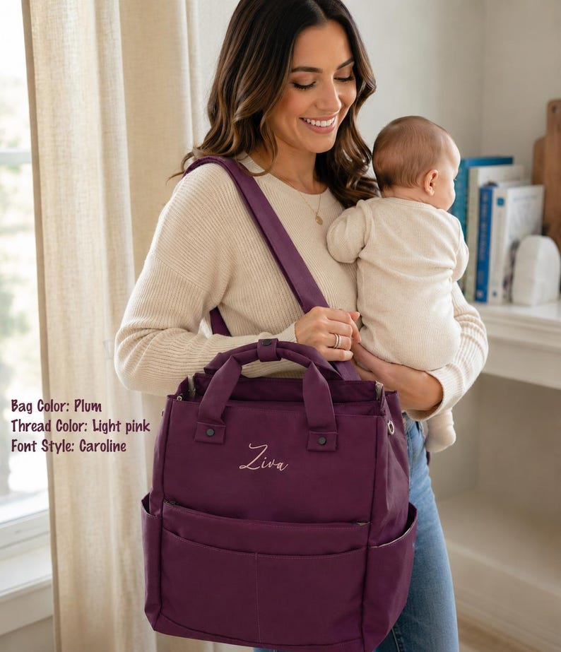 May include: Plum-colored diaper bag with light pink embroidered name "Ziva". The bag has multiple pockets and a top handle, and is carried over the shoulder. The bag is a stylish accessory for parents.