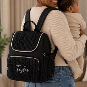May include: Black quilted backpack with gold-colored trim and zippers. The backpack has a top handle, adjustable straps, and a front pocket. The name "Taylor" is embroidered on the front. A stylish and functional bag for everyday use.