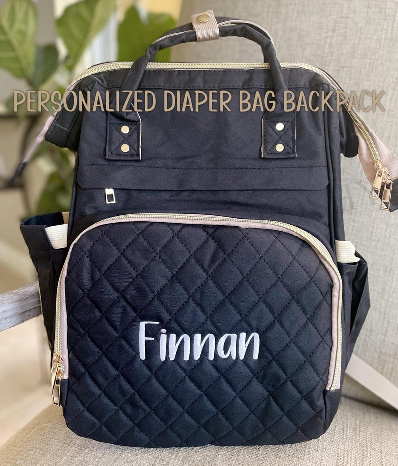 May include: Black diaper bag backpack with a quilted front pocket embroidered with the name "Finnan" in white. The bag has gold zippers, a top handle, and the text "PERSONALIZED DIAPER BAG BACKPACK" across the top.