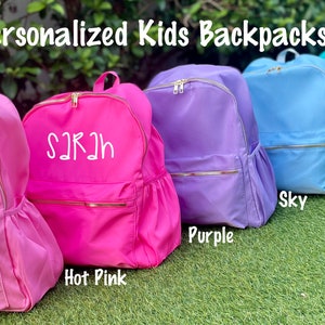 May include: Personalized kids' backpacks in various colors, including light pink, hot pink, purple, sky blue, teal, and multi-colored tie-dye. Each backpack features a personalized name in white lettering.
