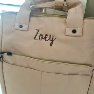 May include: A light pink backpack-style tote bag with a top handle and a front pocket. The name "Zoey" is embroidered in chocolate-colored thread. The bag has black hardware and a zipper closure. The text at the bottom reads "THREAD COLOR: CHOCOLATE FONT: SWEET PEA."