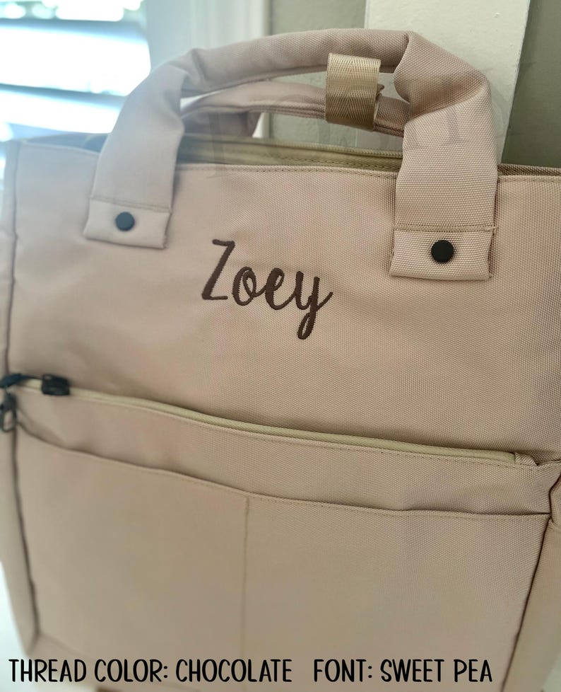 May include: A light pink backpack-style tote bag with a top handle and a front pocket. The name "Zoey" is embroidered in chocolate-colored thread. The bag has black hardware and a zipper closure. The text at the bottom reads "THREAD COLOR: CHOCOLATE FONT: SWEET PEA."