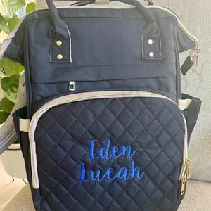 以下が含まれることがあります： 青い刺繍の「Eden Lucah」の文字が入った黒いキルティングおむつバッグ。バッグにはゴールドのジッパー、ライトグレーのトリム、上部のハンドルが付いています。バッグにはフロントポケットとサイドポケットがあります。下部のテキストは「FONT STYLE: SWEET PEA/ THREAD COLOR: MONACO BLUE」です。