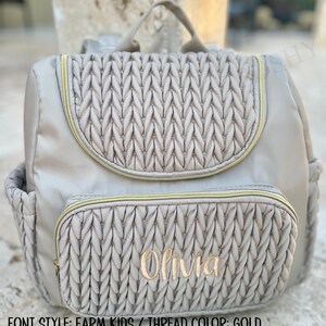 May include: A beige backpack with a quilted flap and front pocket, accented with gold zippers. The name "Olivia" is embroidered in gold thread on the front pocket. Features a top handle and side pockets. The bag is designed for carrying essentials.