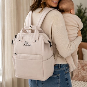 May include: A beige backpack with the name "Blair" in cursive script. The backpack has a top handle, adjustable straps, and multiple pockets. The person in the image is wearing blue jeans and holding a baby.