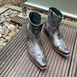 Könnte beinhalten: Ein Paar graue Stiefeletten mit einem strukturierten, schlangenartigen Muster. Die Stiefel haben eine spitze Zehenpartie, einen seitlichen Reißverschluss und eine braune Sohle. Die Stiefel sind ca. 20 cm hoch.