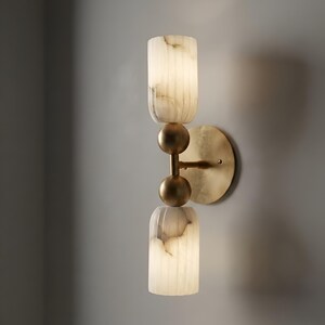 May include: A wall sconce featuring two cylindrical, vertically ribbed white stone shades with natural veining. The shades are separated by two brushed brass spheres and connected to a circular brushed brass wall mounting plate.