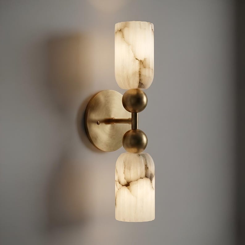 May include: A wall-mounted light fixture featuring a circular brass backplate and a vertical arm. The fixture includes two cylindrical, translucent white stone shades with grey veining, separated by two spherical brass accents in the center.