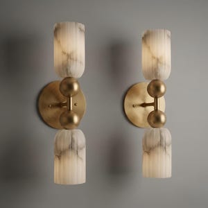 May include: A pair of wall sconces featuring circular brass backplates and central vertical rods with two spherical brass accents. Each fixture holds two cylindrical, vertically fluted, white and grey marbled glass shades that emit a soft glow.