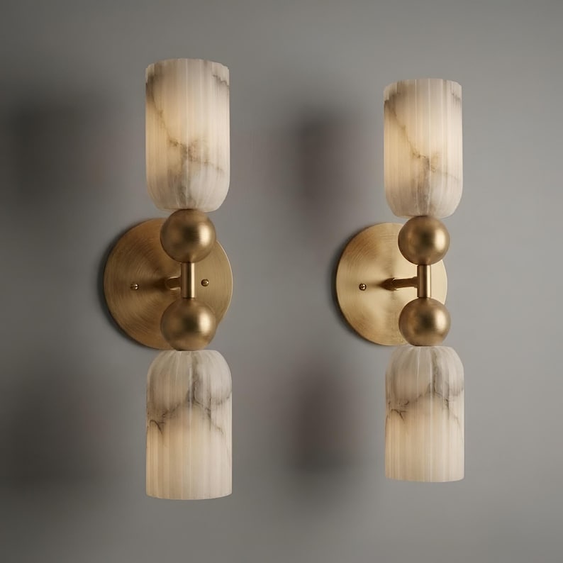 May include: A pair of wall sconces featuring circular brass backplates and central vertical rods with two spherical brass accents. Each fixture holds two cylindrical, vertically fluted, white and grey marbled glass shades that emit a soft glow.