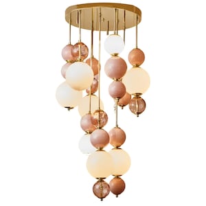 Puede incluir: Una lámpara de araña moderna con una base circular dorada y múltiples hilos colgantes. Cada hilo presenta elementos decorativos esféricos en tonos rosa, melocotón y blanco. Las esferas varían en tamaño y material, creando una lámpara única.