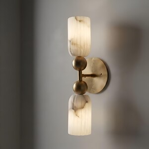 May include: A wall sconce featuring a circular brass backplate and a central vertical rod with two spherical brass accents. Two cylindrical, vertically fluted white stone shades with grey veining are positioned at the top and bottom ends.