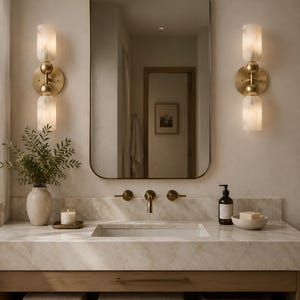 May include: A bathroom vanity featuring a rectangular mirror with rounded corners, two wall-mounted brass sconces with cylindrical white marble shades, a marble countertop, a brass faucet, a ceramic vase, a candle, and a soap dispenser.