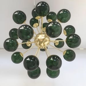 Puede incluir: Una lámpara de araña moderna con múltiples esferas de vidrio verde oscuro que irradian desde una esfera central dorada. Las esferas están unidas a la esfera central por finas varillas doradas, creando un diseño de estrella. La lámpara está suspendida del techo.