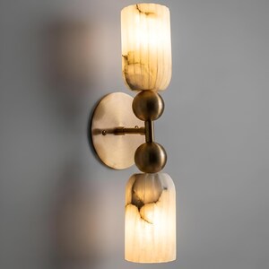 May include: A wall-mounted light fixture featuring a circular brass backplate and a central vertical arm. The arm holds two cylindrical, translucent, cream-colored stone shades with natural veining, separated by two smooth, spherical brass accents.