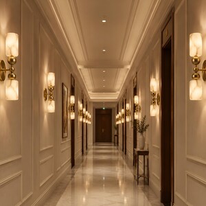 May include: A series of wall-mounted light fixtures featuring two cylindrical, marbled white glass shades separated by a central brass sphere. The fixtures are mounted on circular brass bases and emit a warm, soft glow along a long hallway.