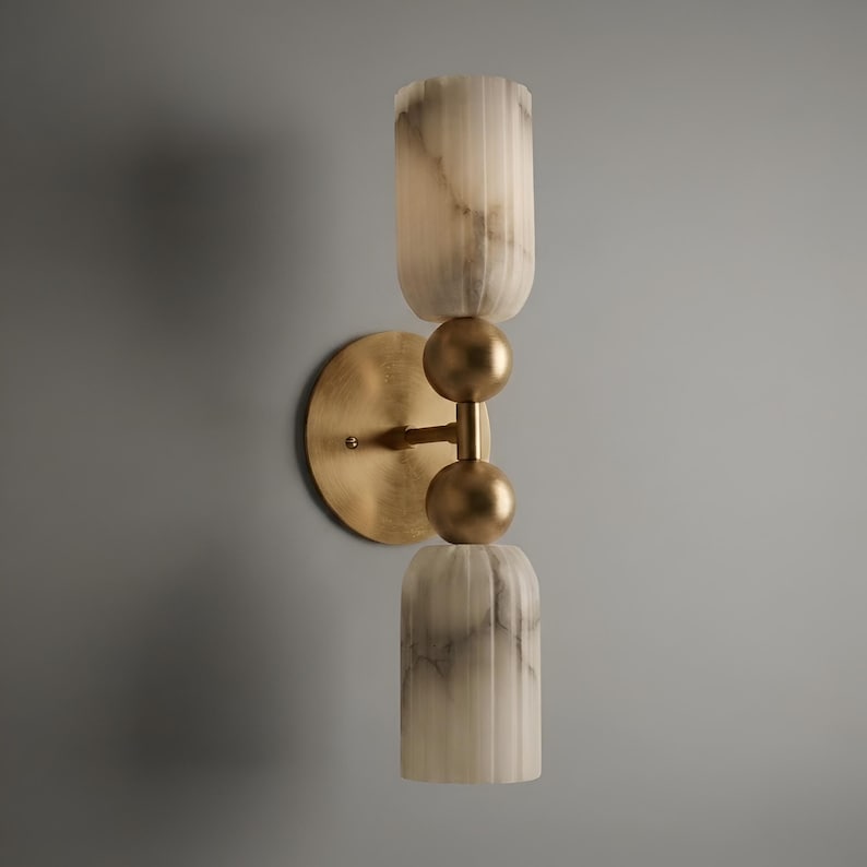 May include: A wall sconce featuring a circular brass backplate and a central vertical rod with two spherical brass accents. The fixture includes two cylindrical, vertically fluted, white and grey marbled glass shades positioned at the top and bottom.