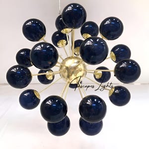 Puede incluir: Una lámpara de araña moderna con esferas azul marino que irradian desde una esfera central dorada. Las esferas están unidas a varillas doradas, creando un diseño de estrella. La lámpara está suspendida del techo, mostrando una lámpara contemporánea.