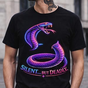May include: Black t-shirt featuring a vibrant graphic of a purple and blue snake with its mouth open, tongue extended. The text "SILENT... BUT DEADLY" is displayed in a gradient font. The snake design has a neon-like glow.