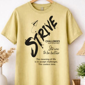 Puede incluir: Una camiseta amarillo claro con la palabra "STRIVE" en letras grandes, negras y estilizadas. Debajo, las palabras "CHALLENGES NEW ADVENTURES" y "Strive to be better". La camiseta también tiene el texto "The meaning of life is to accept challenges. The coolest time."