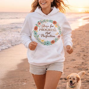 Puede incluir: Sudadera con capucha blanca con un diseño de corona floral y el texto "Strive for Progress Not Perfection". La sudadera tiene un bolsillo delantero y una capucha con cordón. La persona lleva pantalones cortos de color claro y corre en una playa con un perro.
