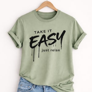Puede incluir: Camiseta verde salvia con mangas enrolladas, colgada de una percha de madera. La camiseta presenta las palabras "TAKE IT EASY" en una fuente negra y audaz, con "just relax" debajo. La camiseta se combina con unos vaqueros azules.