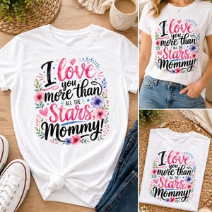 May include: White t-shirt with the text "I love you more than all the stars, Mommy!" in a decorative font, surrounded by floral and leaf accents. The shirt is paired with blue jeans and white sneakers, suggesting casual wear.