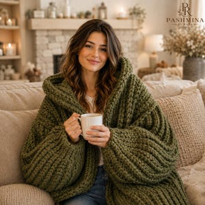 Könnte beinhalten: Eine olivgrüne, grob gestrickte Kapuzenstrickjacke mit langen Ärmeln und einer dicken Rippenstruktur. Das Kleidungsstück verfügt über eine große Kapuze und ein offenes Vorderteil, getragen über einem schlichten weißen Oberteil mit einer weißen Tasse.