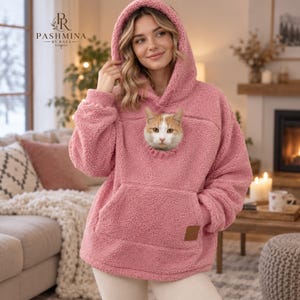 Może przedstawiać: Bluza z kapturem z polaru typu sherpa w kolorze przybrudzonego różu, wyposażona w dużą kieszeń typu kangurka z okrągłym otworem na klatce piersiowej. Przez otwór wygląda rudo-biały kot. Odzież posiada kaptur oraz małą brązową naszywkę w dolnej części.
