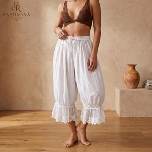 May include: White calf-length bloomers featuring a drawstring elastic waistband and delicate lace trim at the hem. The garment is paired with a brown lace bralette. The text PASHMINA BY RAZA is visible in the upper left corner of the image.