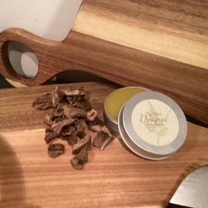 Könnte beinhalten: Zwei Holzschneidebretter, eines mit Griff. Ein kleiner Haufen getrockneter, brauner Stücke liegt auf dem Brett. Eine kleine, silberne Dose mit dem Wort "Designed" befindet sich neben einer kleinen Dose mit gelbem Balsam.