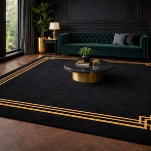 May include: A large black rug with a gold geometric border. The rug is on a hardwood floor in a room with a green velvet sofa, gold accents, and a round coffee table. The rug's dimensions are approximately 240 cm x 300 cm.