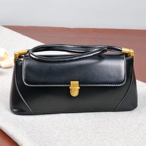 May include: A black handbag with a rectangular shape and a gold-coloured clasp. The bag has a handle and gold accents on the sides. The bag is on a light grey surface.