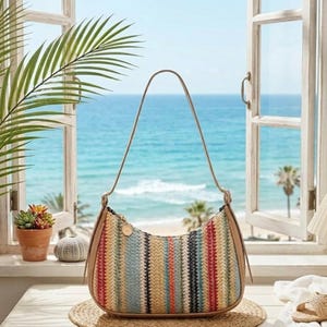 May include: A crescent-shaped shoulder bag featuring a woven texture with vertical stripes in shades of tan, blue, red, and cream. The bag has a smooth tan trim along the edges and a single thin shoulder strap attached with gold-toned hardware.