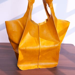 K&ouml;nnte beinhalten: Eine senfgelbe Ledertasche mit zwei Schulterriemen. Die Tasche hat ein gro&szlig;es, offenes Design mit rechteckiger Basis und einer weichen, unstrukturierten Form. Das Leder wirkt glatt und hat einen leichten Glanz, was auf eine hochwertige Verarbeitung hindeutet.