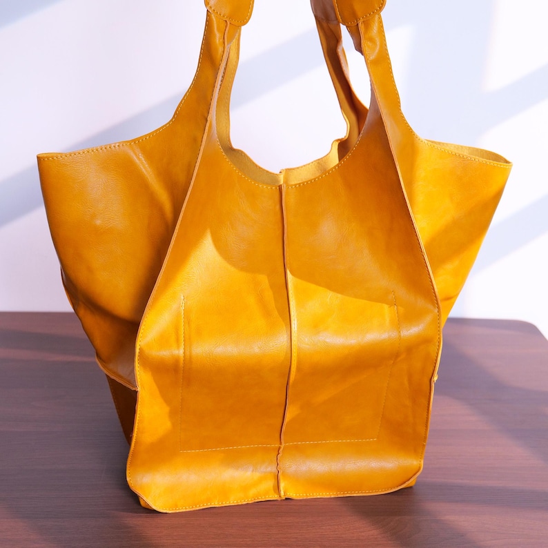 K&ouml;nnte beinhalten: Eine senfgelbe Ledertasche mit zwei Schulterriemen. Die Tasche hat ein gro&szlig;es, offenes Design mit rechteckiger Basis und einer weichen, unstrukturierten Form. Das Leder wirkt glatt und hat einen leichten Glanz, was auf eine hochwertige Verarbeitung hindeutet.