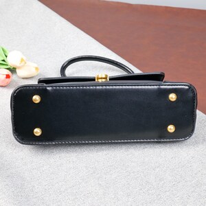 May include: A black leather handbag with a top handle and gold-coloured hardware. The bag has a rectangular shape with rounded corners and four gold-coloured feet on the bottom. The clasp is gold-coloured.