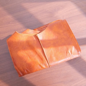 K&ouml;nnte beinhalten: Gefaltete, orangebraune Ledertasche mit strukturiertem Design. Die Tasche hat eine glatte Textur und sichtbare Nahtdetails. Das Bild ist auf einer hellbraunen Holzoberfl&auml;che mit nat&uuml;rlichen Schatten aufgenommen.