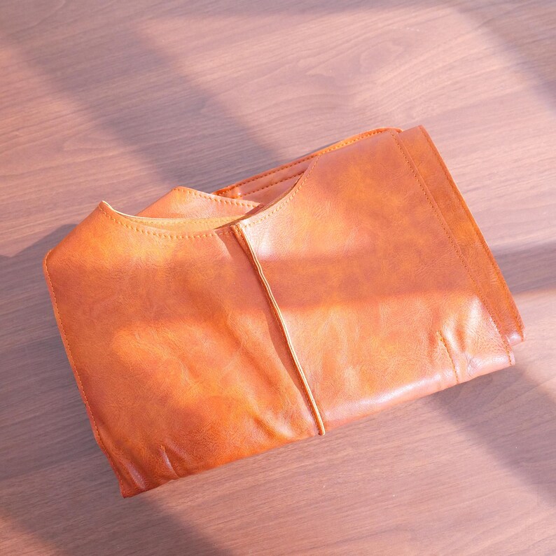 K&ouml;nnte beinhalten: Gefaltete, orangebraune Ledertasche mit strukturiertem Design. Die Tasche hat eine glatte Textur und sichtbare Nahtdetails. Das Bild ist auf einer hellbraunen Holzoberfl&auml;che mit nat&uuml;rlichen Schatten aufgenommen.