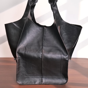 K&ouml;nnte beinhalten: Eine schwarze Tragetasche aus leder&auml;hnlichem Material. Die Tasche hat zwei Griffe und ein gro&szlig;es offenes Fach. Sie hat eine rechteckige Tasche auf der Vorderseite. Die Tasche steht auf einer Holzoberfl&auml;che.