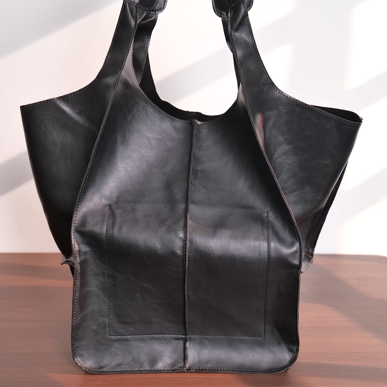 K&ouml;nnte beinhalten: Eine schwarze Tragetasche aus leder&auml;hnlichem Material. Die Tasche hat zwei Griffe und ein gro&szlig;es offenes Fach. Sie hat eine rechteckige Tasche auf der Vorderseite. Die Tasche steht auf einer Holzoberfl&auml;che.
