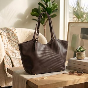May include: A dark brown leather tote bag with two long shoulder straps featuring adjustable gold-tone buckles. The front of the bag is embossed with the word JAMES in small capital letters. The bag has a wide opening and a structured base.