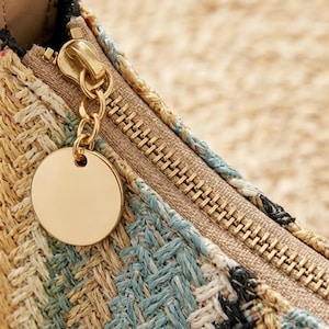 May include: A woven bag featuring a textured pattern in shades of tan, light blue, and black. The bag includes a gold-toned metal zipper closure with a matching circular metal charm attached to the zipper pull, set against a beige fabric lining.