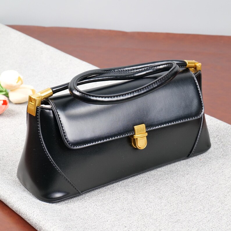 May include: A black handbag with a curved handle and gold-coloured hardware. The rectangular bag has a flap closure and a structured design. The bag is made of a shiny material and sits on a light grey surface.