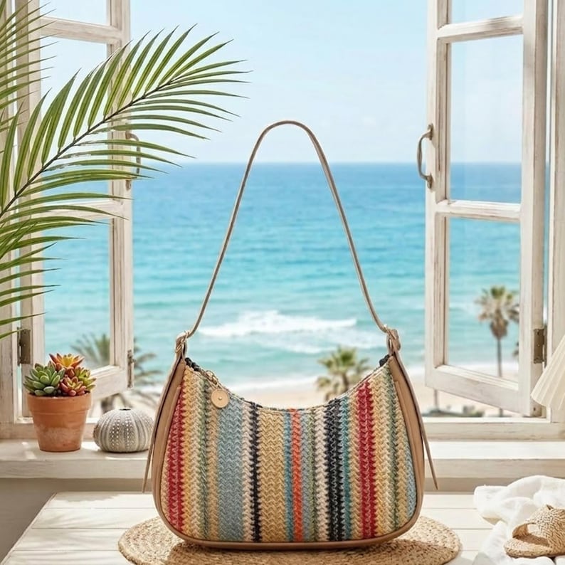 May include: A crescent-shaped shoulder bag featuring a woven texture with vertical stripes in shades of tan, blue, red, and cream. The bag has a smooth tan trim along the edges and a single thin shoulder strap attached with gold-toned hardware.