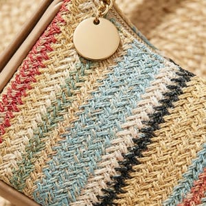 May include: A woven straw pouch featuring a diagonal striped pattern in shades of tan, red, green, light blue, and black. The pouch includes a smooth, round gold-toned metal zipper pull attached to a small chain link on the top edge.