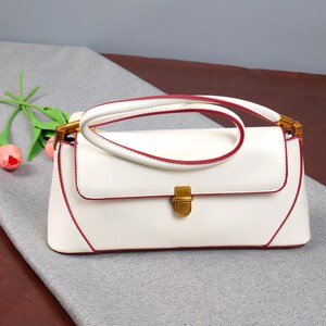 May include: A white handbag with a rectangular shape and a curved handle. The bag has red trim along the edges and handle, with gold-coloured hardware on the clasp and handle attachments. The bag is on a grey surface.